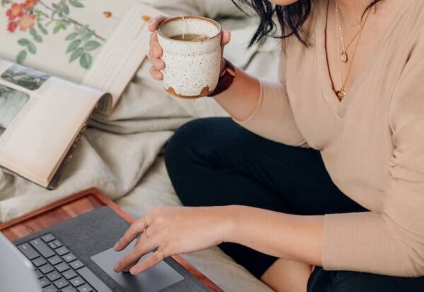 woman in beige long sleeve shirt and black pants sitting on bed using a Microsoft Surface Laptop 3